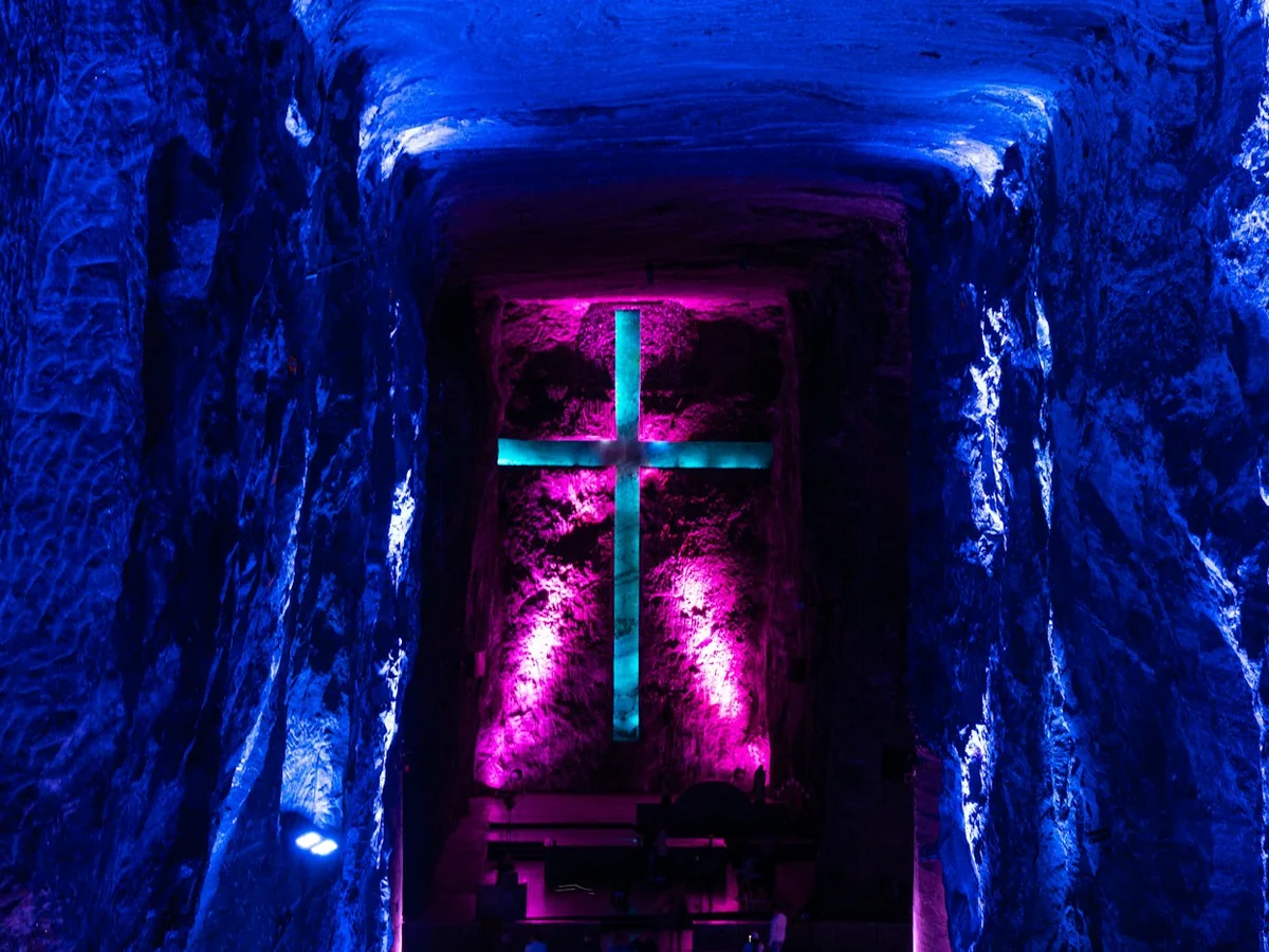 A teal cross lit against a magenta-lit salt wall at the end of a blue-lit tunnel inside the Salt Cathedral